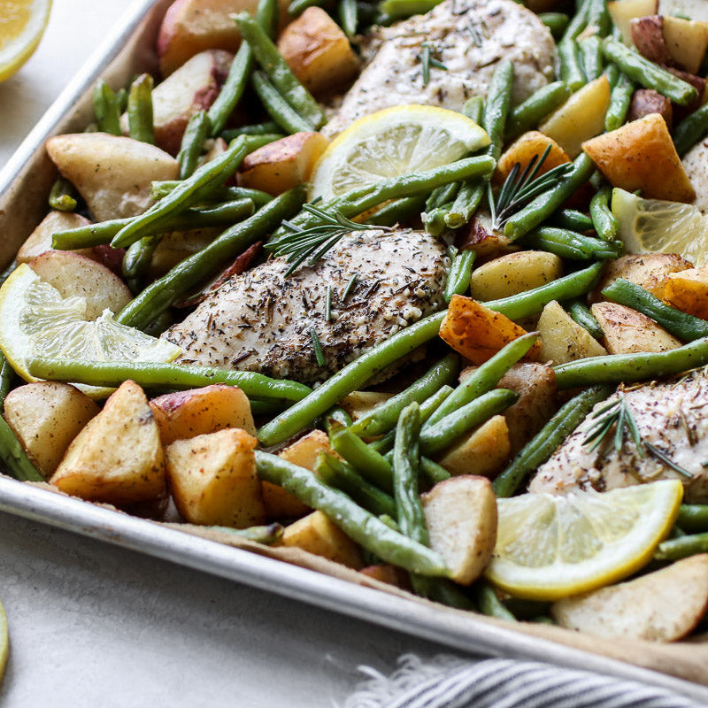Sheet pan with roasted chicken, green beans, and potatoes garnished with lemon slices and rosemary.